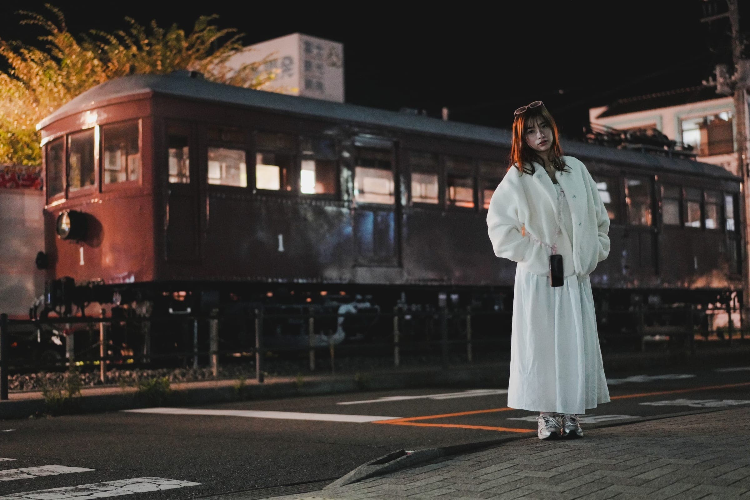 A woman in white standing beside a vintage red train carriage at dusk